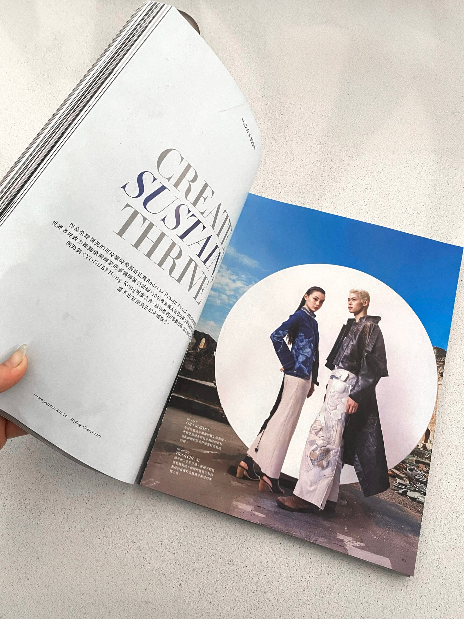 Hong Kong Vogue magazine laying on a cream marble bend. A hand opening the magazine revealing featuring an image of two models wearing the design work of two up and coming sustainable designers.